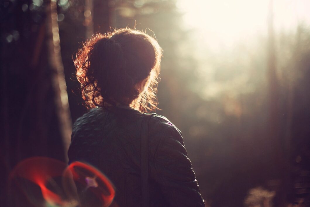 Silhouette of a woman standing in a forest, looking into the sunlight with a contemplative expression.