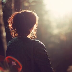 Silhouette of a woman standing in a forest, looking into the sunlight with a contemplative expression.