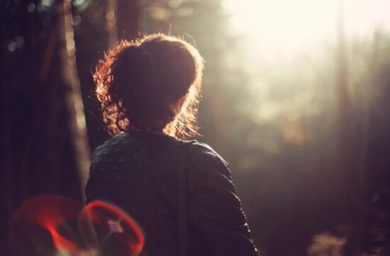 Silhouette of a woman standing in a forest, looking into the sunlight with a contemplative expression.